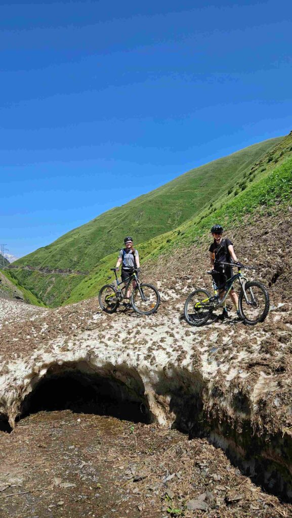 Two mountain bikers crossing a summer snow arch over a mountain stream, Gudamakari, Georgia